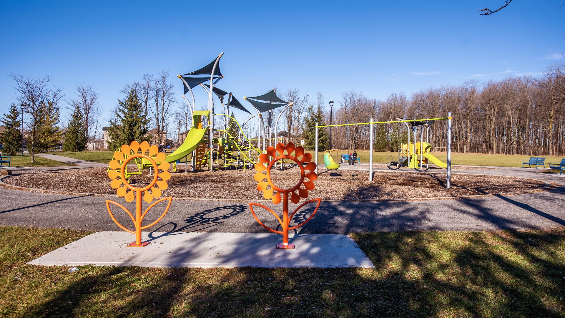 Bike racks in the shape of two sunflowers in the foreground; behind is a children's playground with slides, swings, climbing structures, and other elements