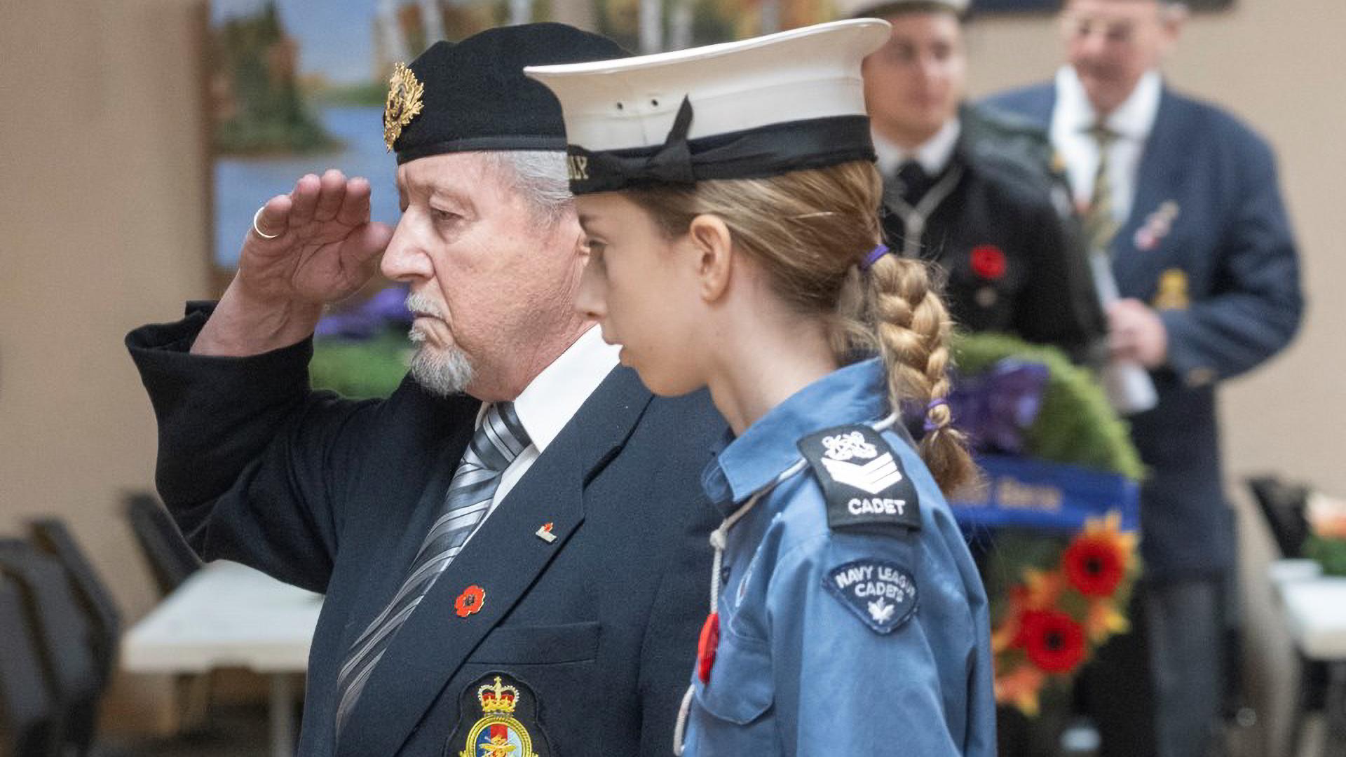 Man giving a solid salute in front of the rows of wreaths alongside young Navy Cadet