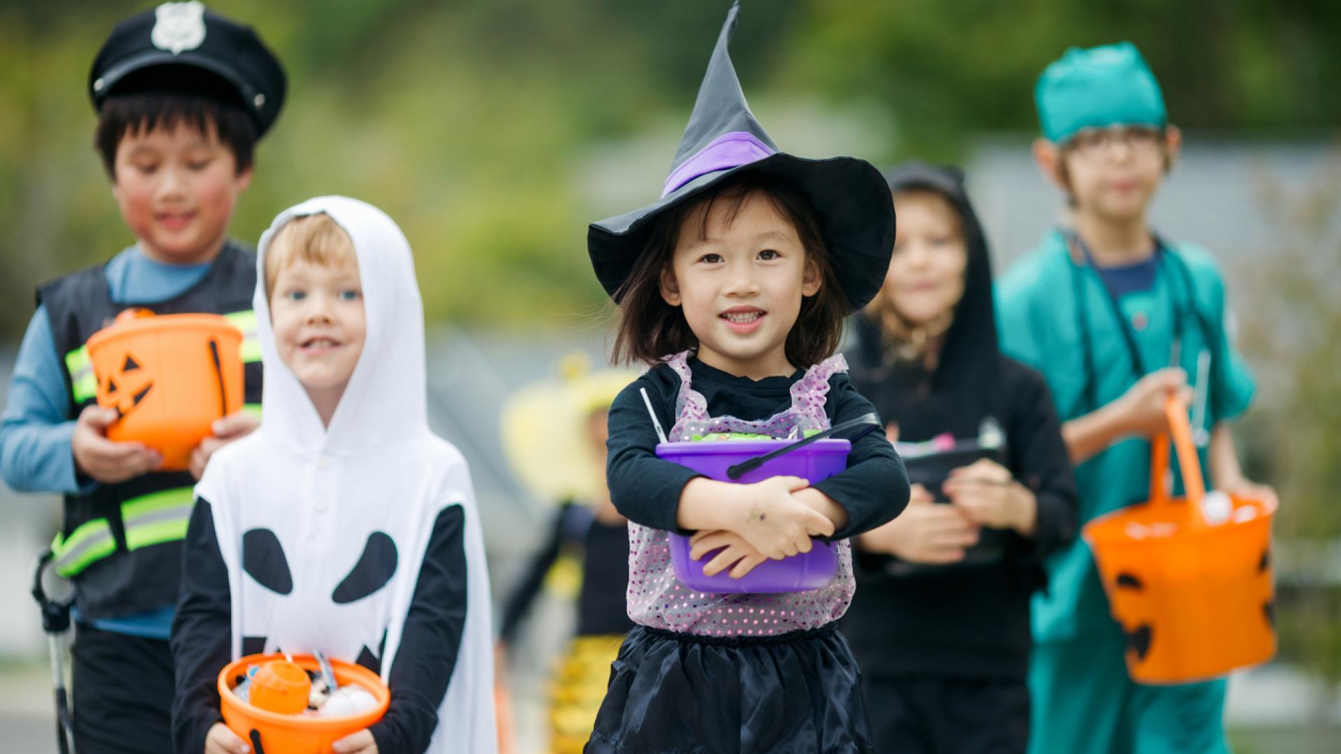 A group of young kids trick or treating for Halloween. They wear assorted traditional costumes, carrying Jack-o'-lantern buckets filled with candy.