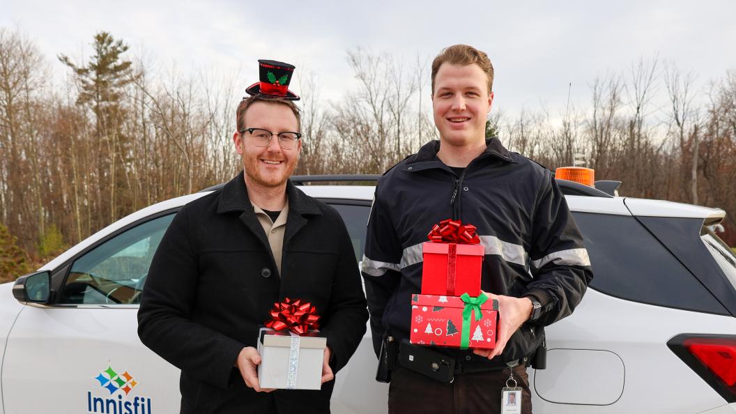 Two Municipal Law Enforcement Officers holding presents outside by-law vehicle in festive cheer