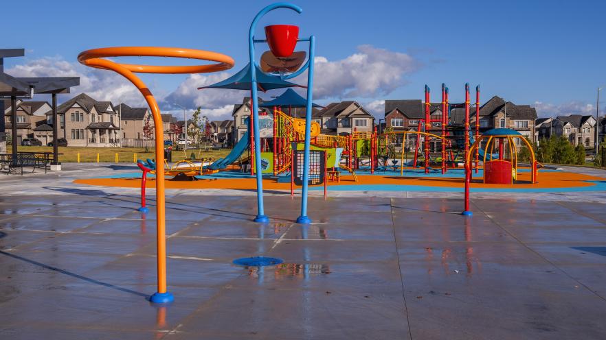 Accessible splash pad with colourful water play features, including spray loops and a tipping bucket, beside a playground in a residential neighbourhood.